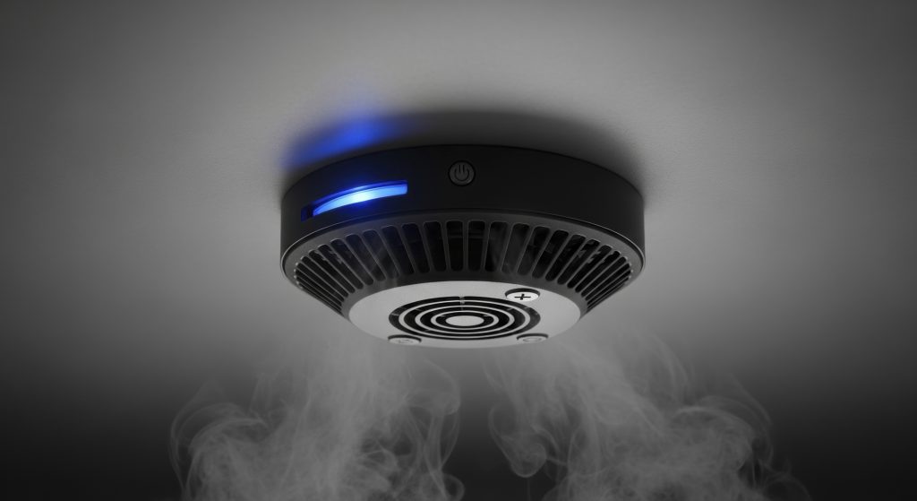 Modern smoke detector with blue light detecting smoke on a grey ceiling for home safety.